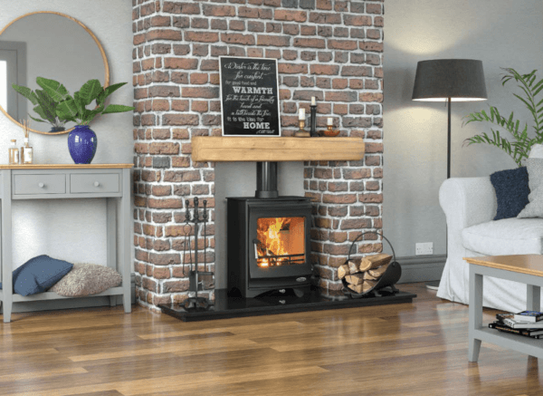 Stunning black stove with fire lit behind large viewing window and matching blaack pipe work. Set on a black granite hearth with rustic timber mantle and brick feature wall
